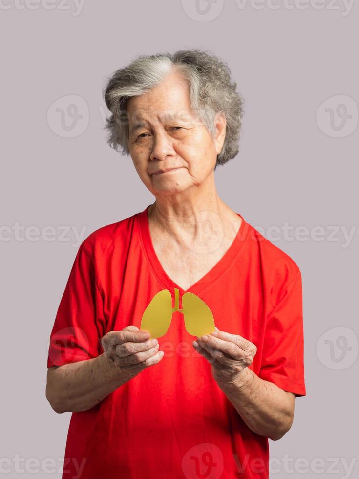 A senior woman holding a lungs shape made from yellow paper while standing on a red background photo