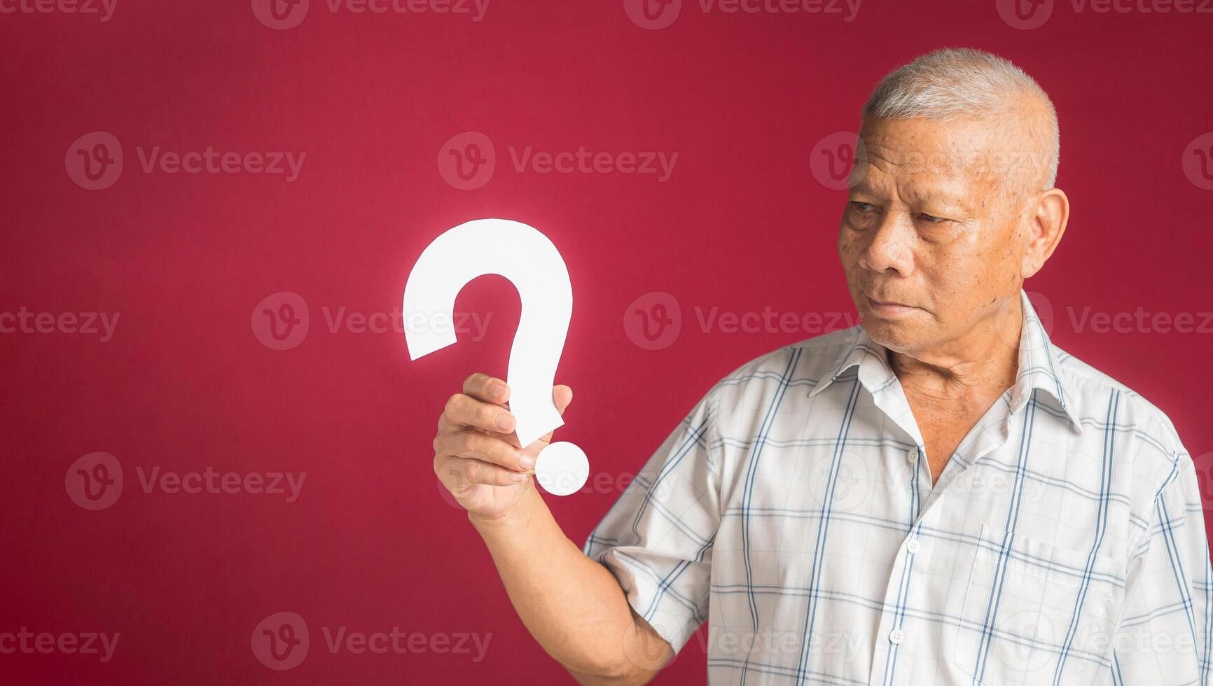 Portrait of a senior man holding and looking at a white question mark while standing with a red background photo