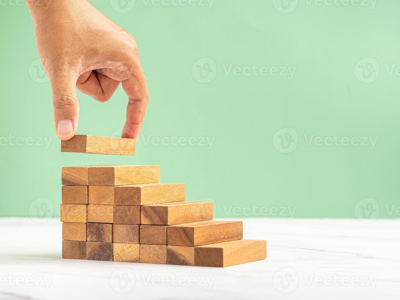Close-up of hand holding a wooden block arranging to stack for development as step stair on a marble floor over a green background photo