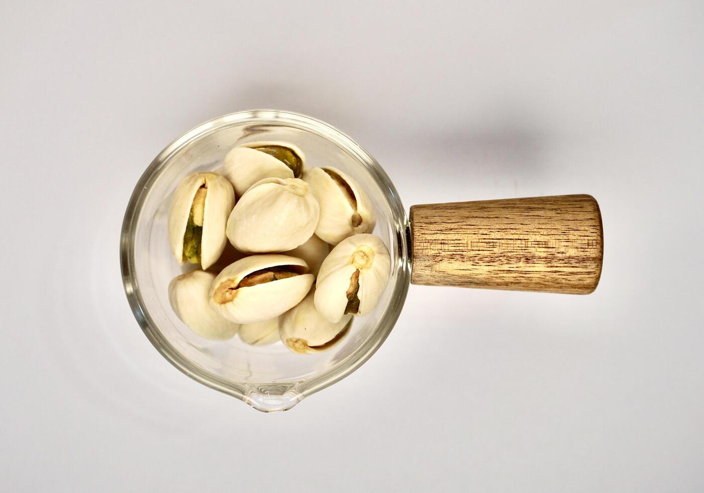 Pile of pistachio nuts in small glassware pot cup with wooden handle isolated on horizontal white background from top angle view. photo