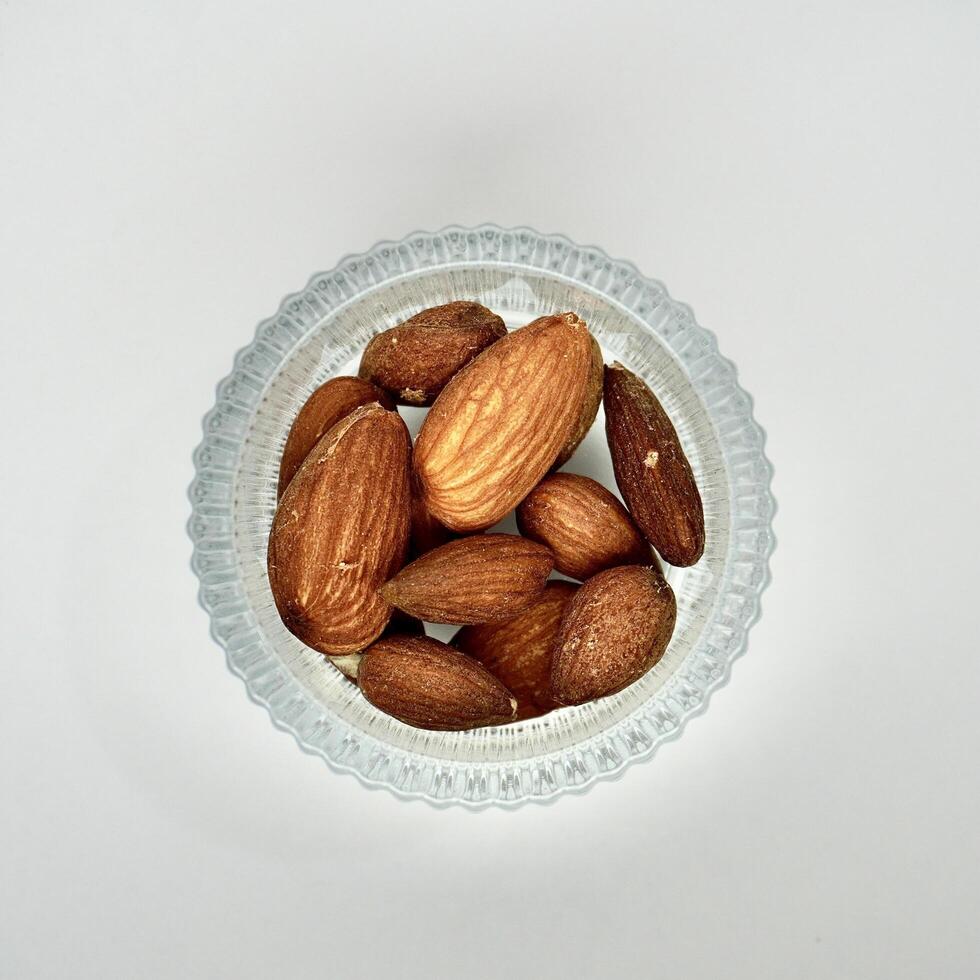 Pile of brown almonds in round glass bowl isolated on square white background from top angle view. photo