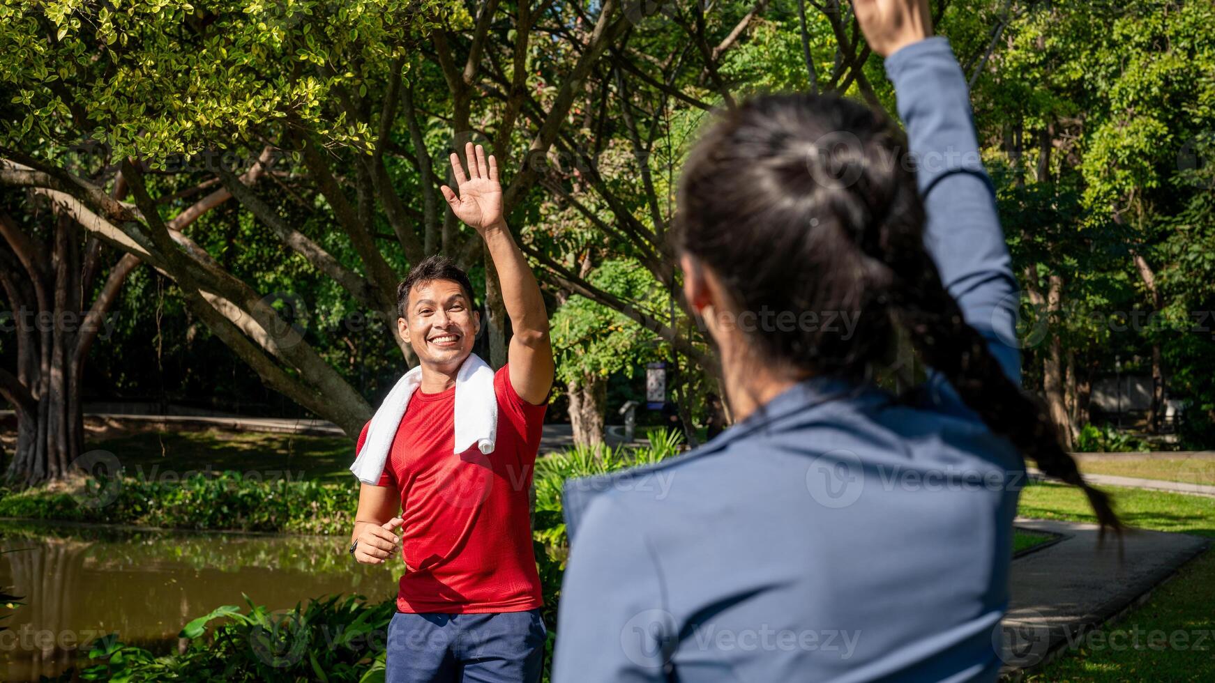 Cheerful Asian man waves and greets his friend as they bump into each other while jogging in a park. photo