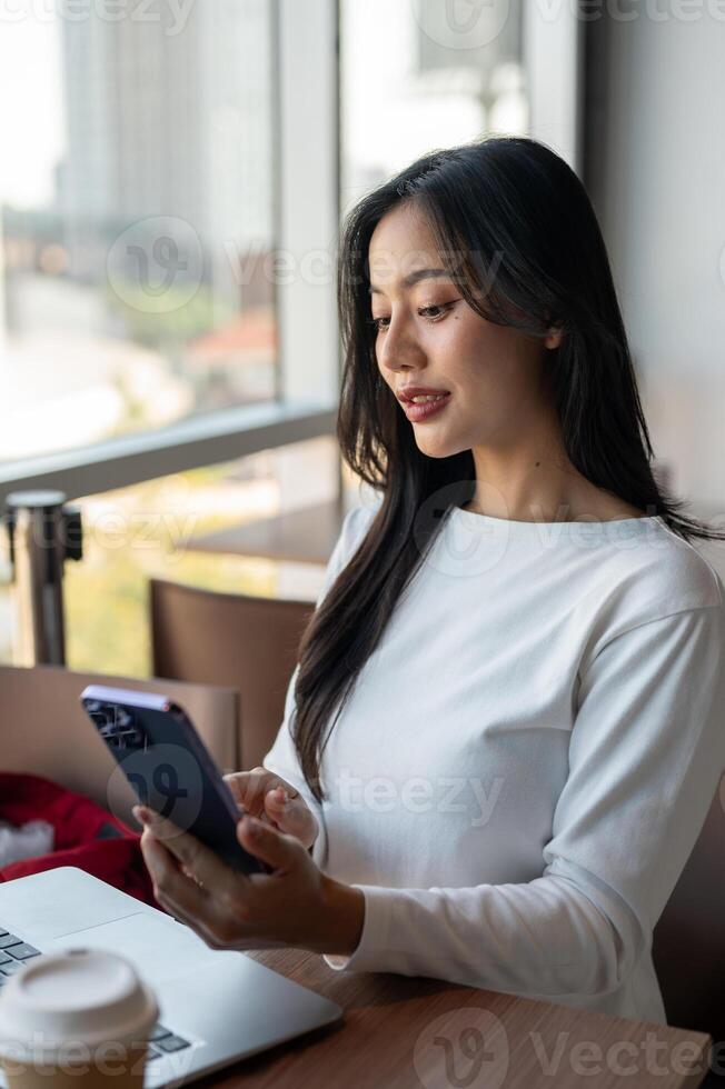 Attractive Asian woman reading messages on her smartphone while working remotely from a coffee shop. photo