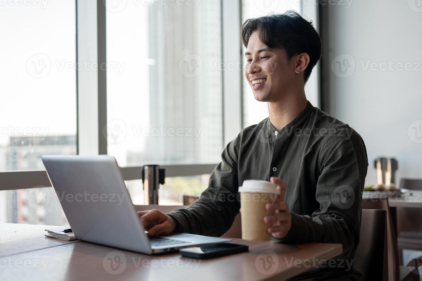 A positive Asian businessman enjoying a hot drink while working on his laptop, working remotely. photo