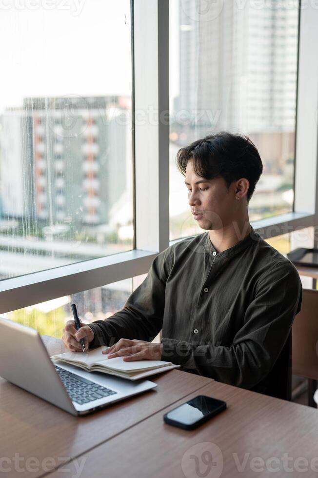 A focused businessman working in his office, writing in a book while sitting in front of a computer. photo