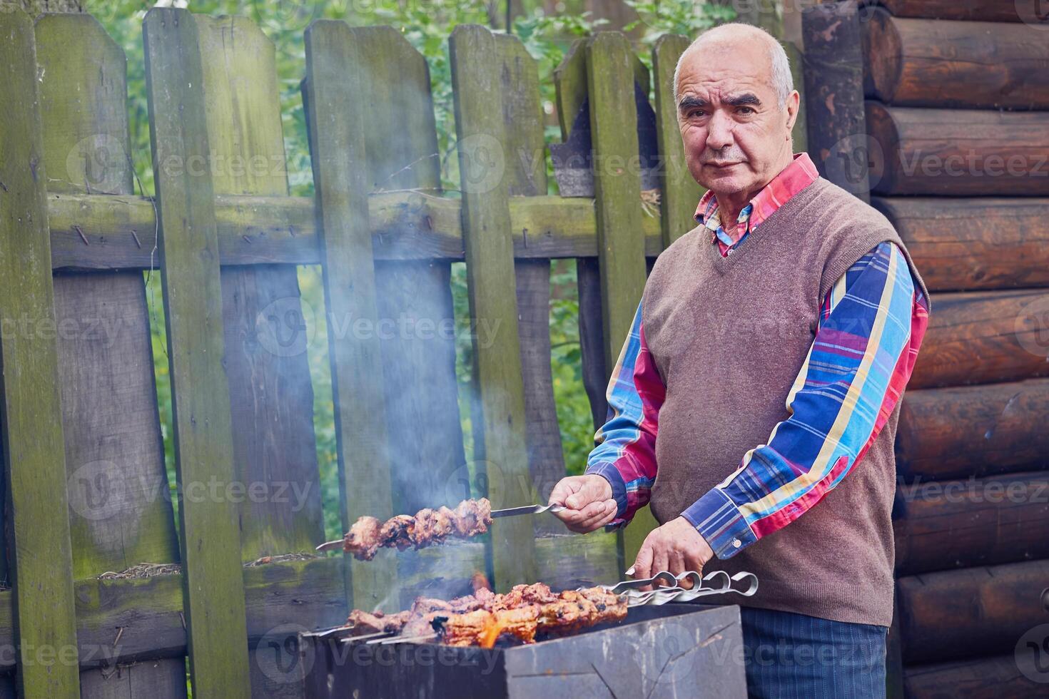 Elderly cheerful pensioner prepares meat for barbecue for cooking on grill fire photo