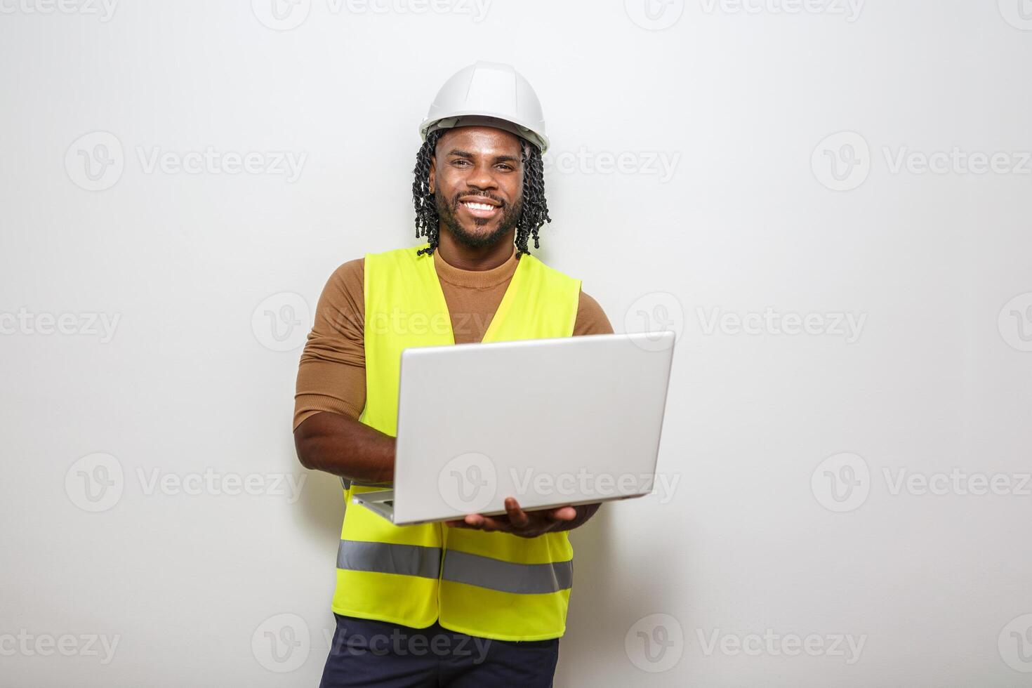 Construction worker with laptop smiles while standing against a plain background in a safety vest photo