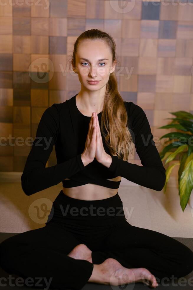 An athletic girl in black sportswear sits in lotus pose at a yoga class in a fitness room among green indoor plants photo