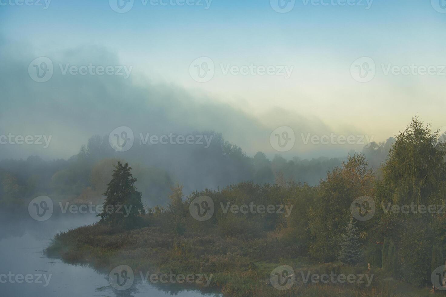 Misty morning landscape featuring a tranquil river with trees reflected in the calm water photo