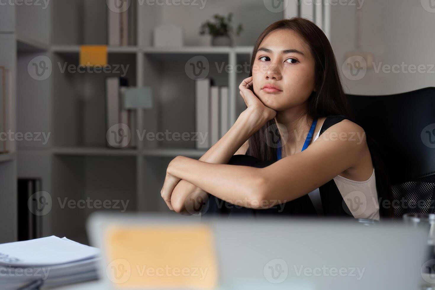 Bored Woman at Work Desk Staring into Distance in Modern Office Setting with Papers and Laptop photo