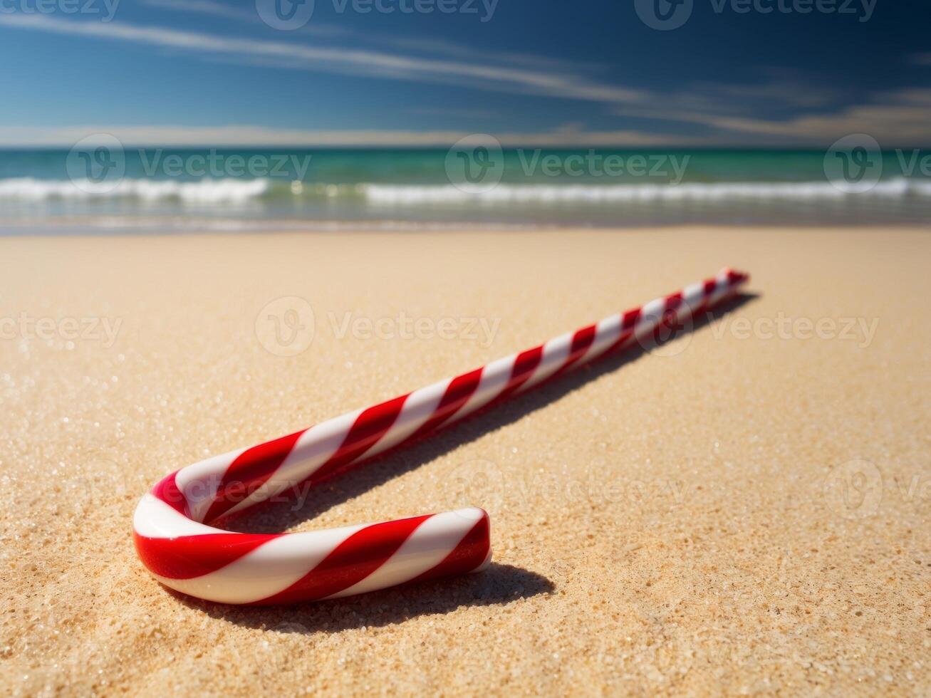 A festive candy cane stands out on sunlit beach sand with gentle waves and a bright blue sky in the background. photo