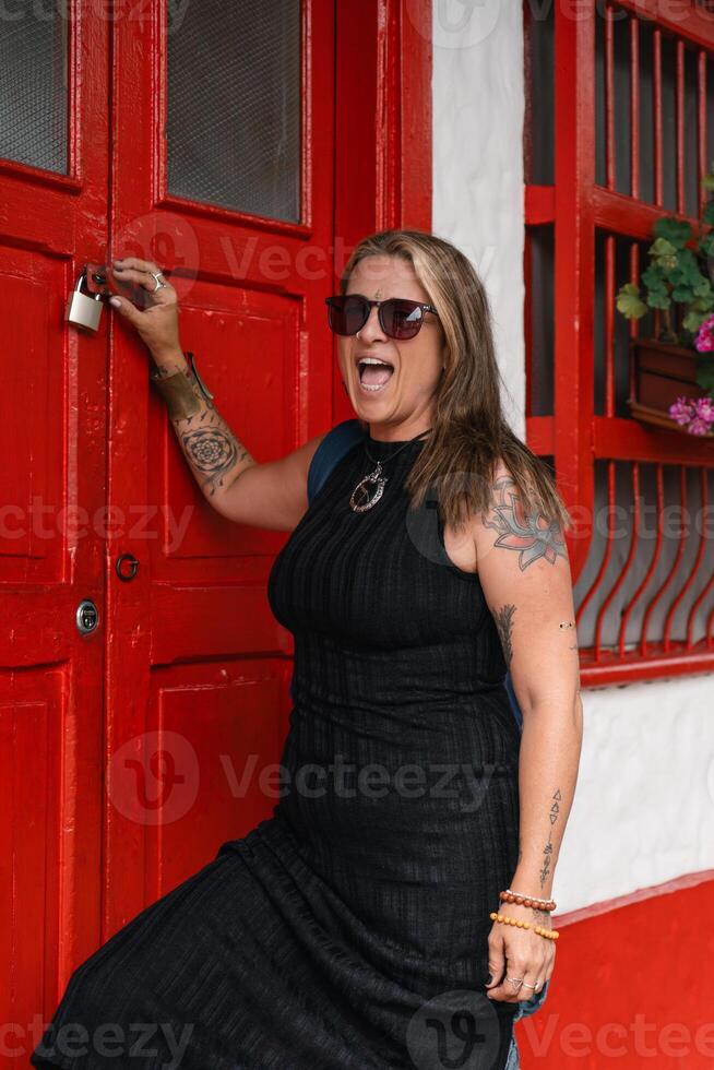 Smiling woman with tattoos and sunglasses posing by a vibrant red door in a charming urban setting photo
