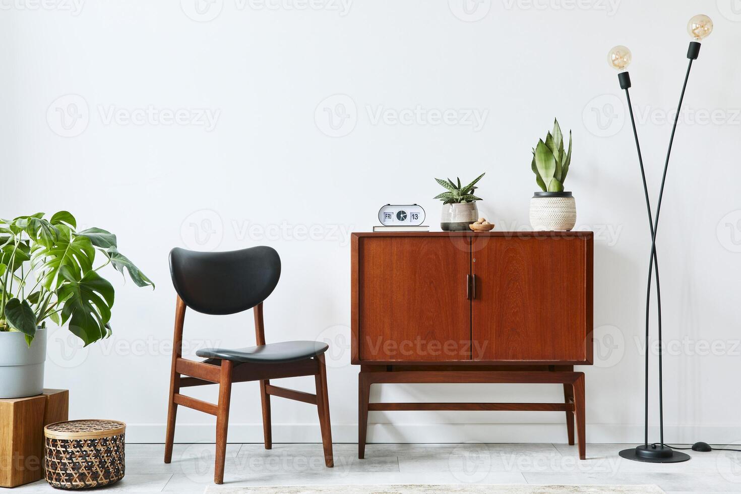 Stylish interior design of living room with wooden retro commode, chair, few plants, rattan basket, decoration, lamp, clock and elegant personal accessories. Copy space white wall. Template. photo