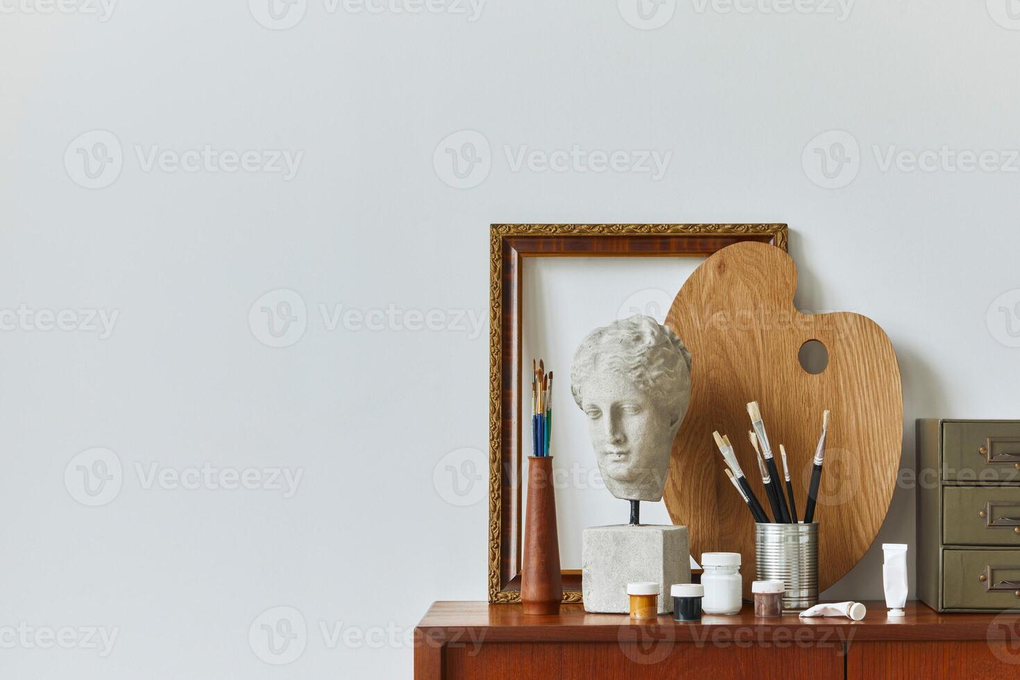 Stylish composition of artist workspace room with design retro teak commode, mock up poster frame, book, decoration and painting accessories. Template. photo