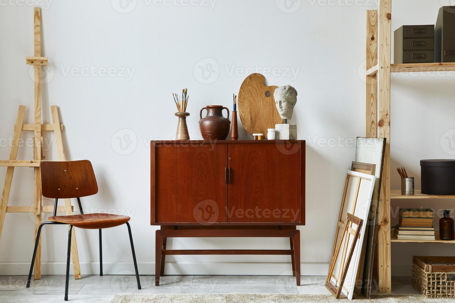 Stylish composition of artist workspace with design retro teak commode, chair, bookcase, poster frames, easel, decoration and elegant personal accessories. Template. photo