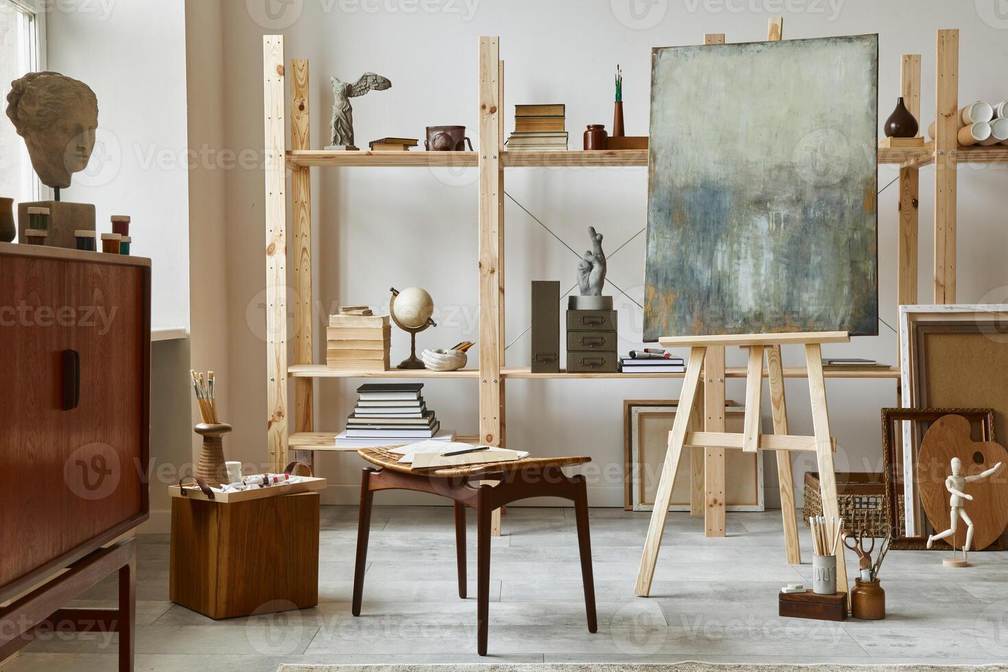 Unique artist workspace interior with stylish teak commode, wooden easel, bookcase, artworks, painting accessories, decoration and elegant personal stuff. Modern work room for artist. Template. photo