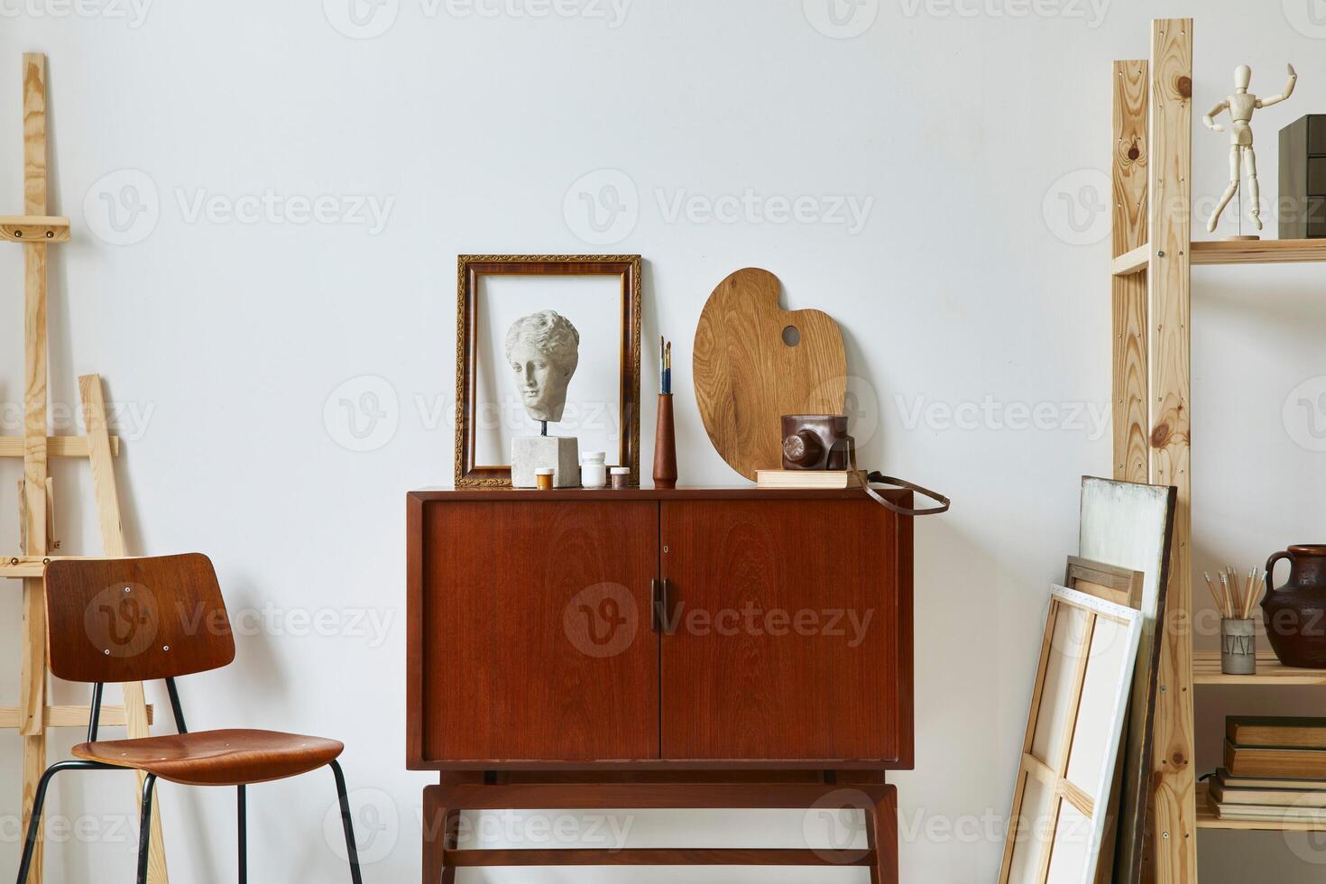 Stylish composition of artist workspace with design retro teak commode, chair, bookcase, poster frames, easel, decoration and elegant personal accessories. Template. photo