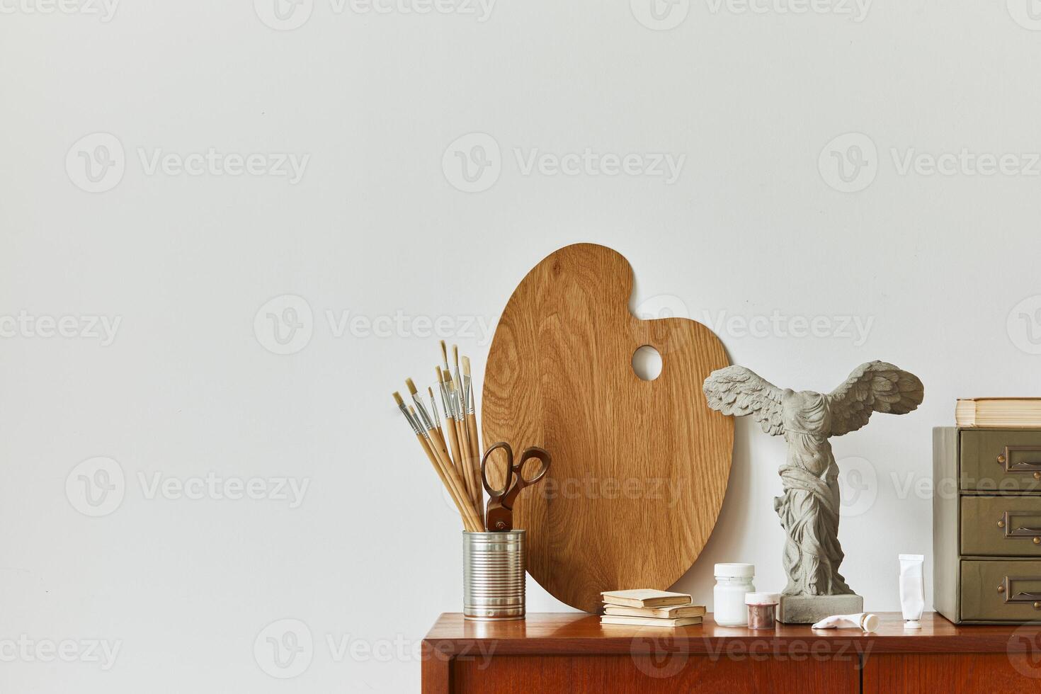 Stylish composition of artist workspace room with design retro teak commode, mock up poster frame, book, decoration and painting accessories. Template. photo