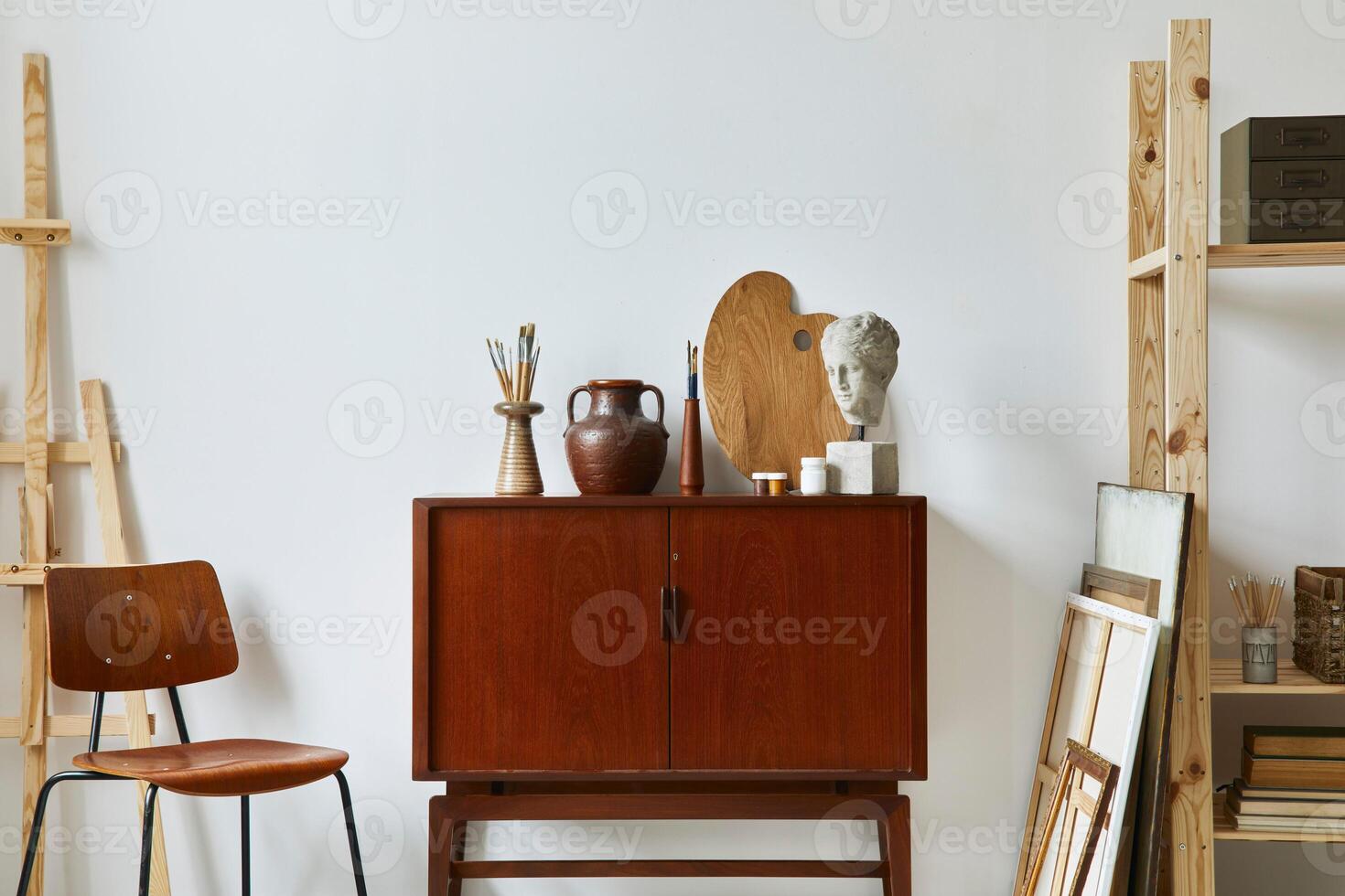 Stylish composition of artist workspace with design retro teak commode, chair, bookcase, poster frames, easel, decoration and elegant personal accessories. Template. photo