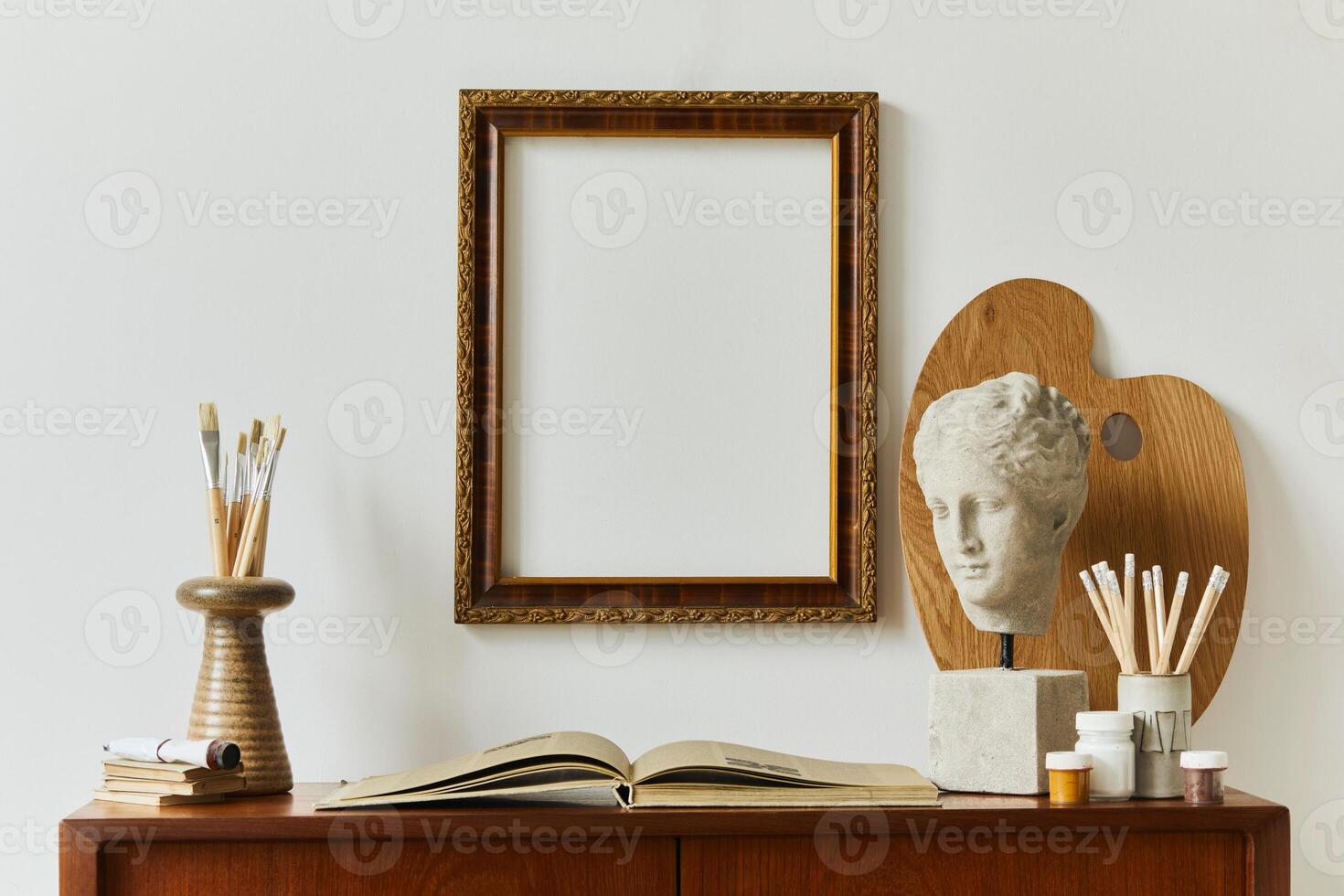 Stylish composition of artist workspace room with design retro teak commode, mock up poster frame, book, decoration and painting accessories. Template. photo