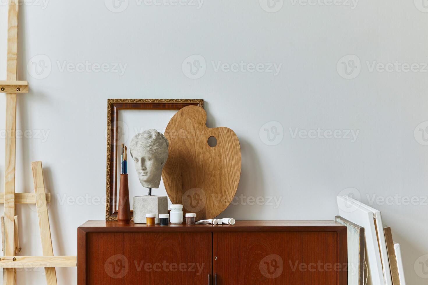 Stylish composition of artist workspace room with design retro teak commode, mock up poster frame, book, decoration and painting accessories. Template. photo