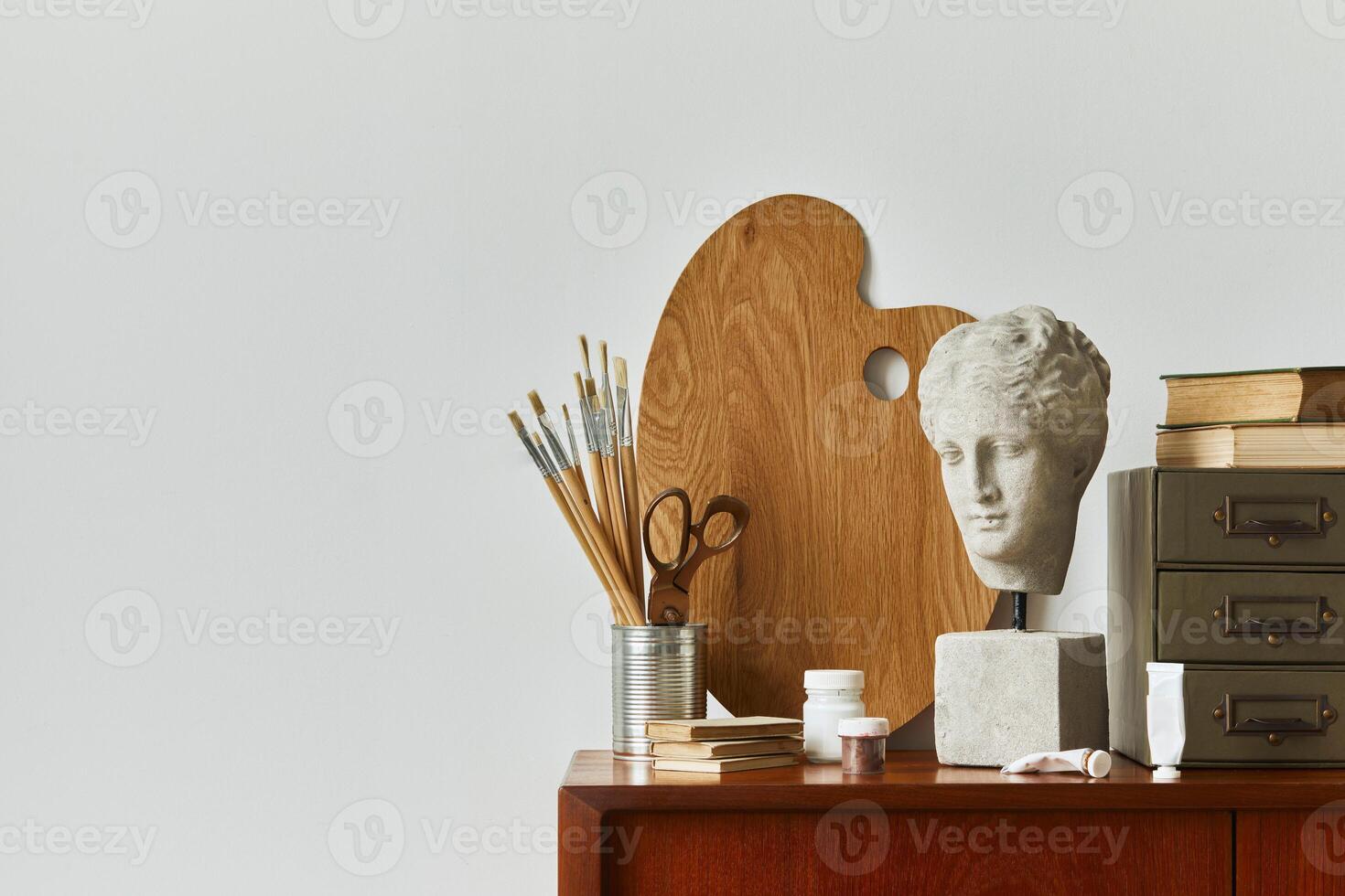 Stylish composition of artist workspace room with design retro teak commode, mock up poster frame, book, decoration and painting accessories. Template. photo