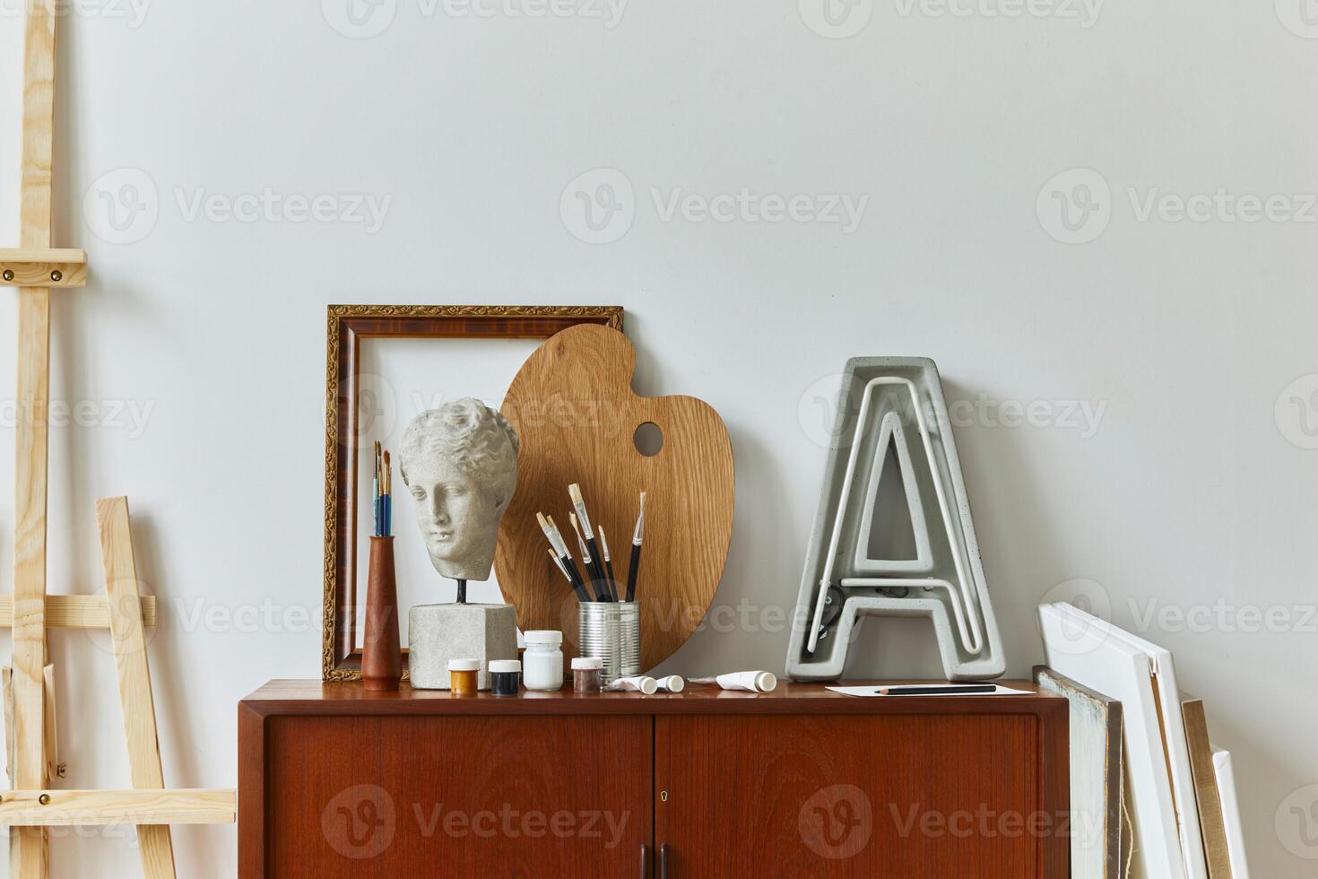 Stylish composition of artist workspace room with design retro teak commode, poster frames, easel, book, decoration and painting accessories. Template. photo