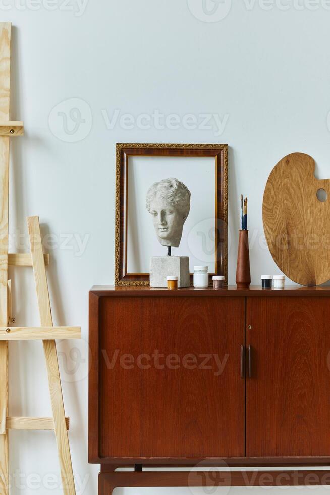 Stylish composition of artist workspace room with design retro teak commode, mock up poster frame, book, decoration and painting accessories. Template. photo