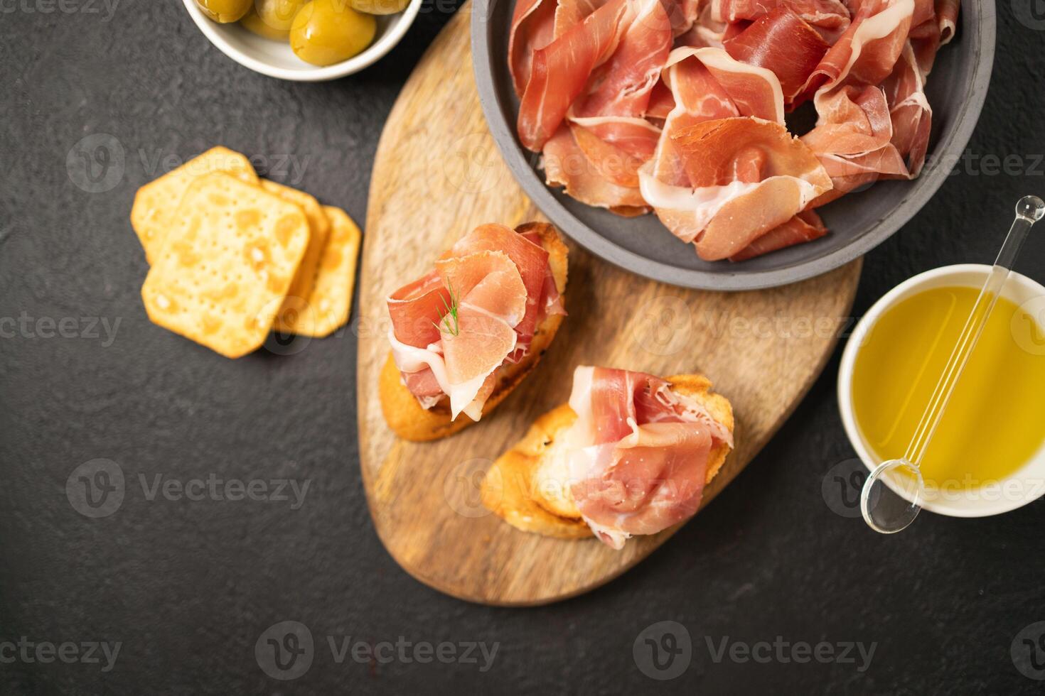 Italian sandwiches bruschetta with olives, bran bread, cherry tomatoes, prosciutto on wooden cutting board, dark black background surface photo