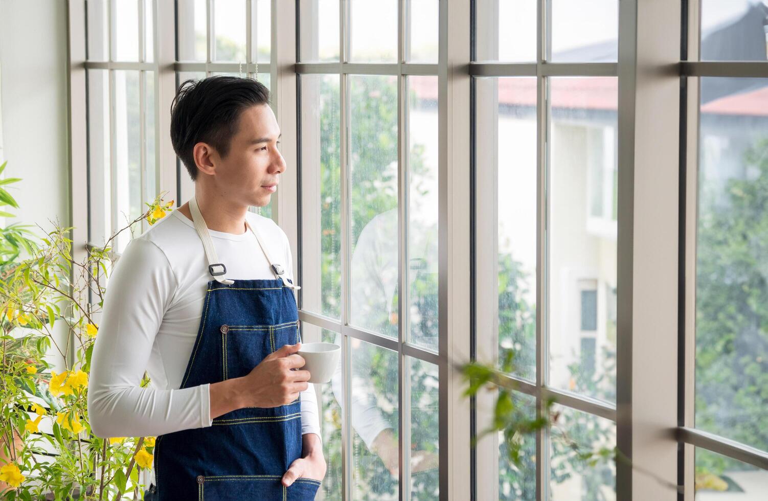 Portrait gardener adult young Asian man one person handsome standing think look and Smile relax happy hand holding drinking cup coffee in room cafe with glass window and green tree in morning time. photo