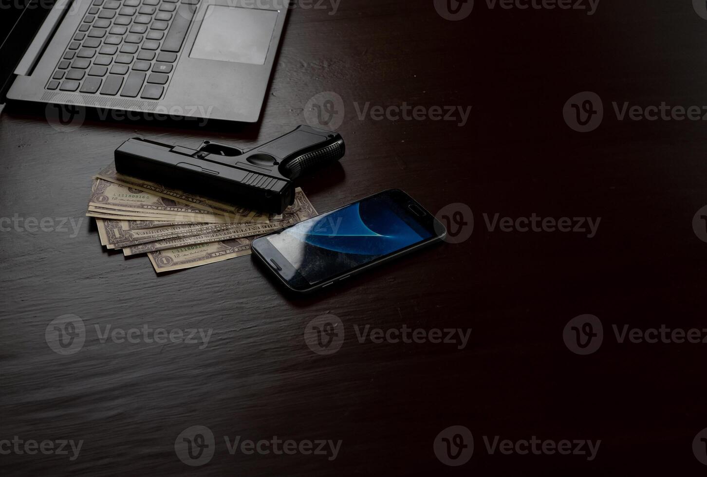 Computers, mobile phone, money, guns, and other devices that hackers use to steal or hijack information to trick victims into taking money. laying on wooden table in dark room in mysterious apartment. photo