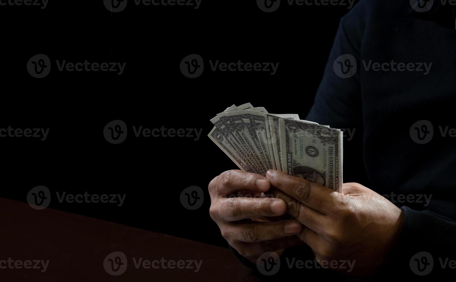 Hands of a man wearing a black shirt, sitting on a chair and a table, is a thief, holding money, counting the amount obtained from hijacking or robbing, in a dark background photo