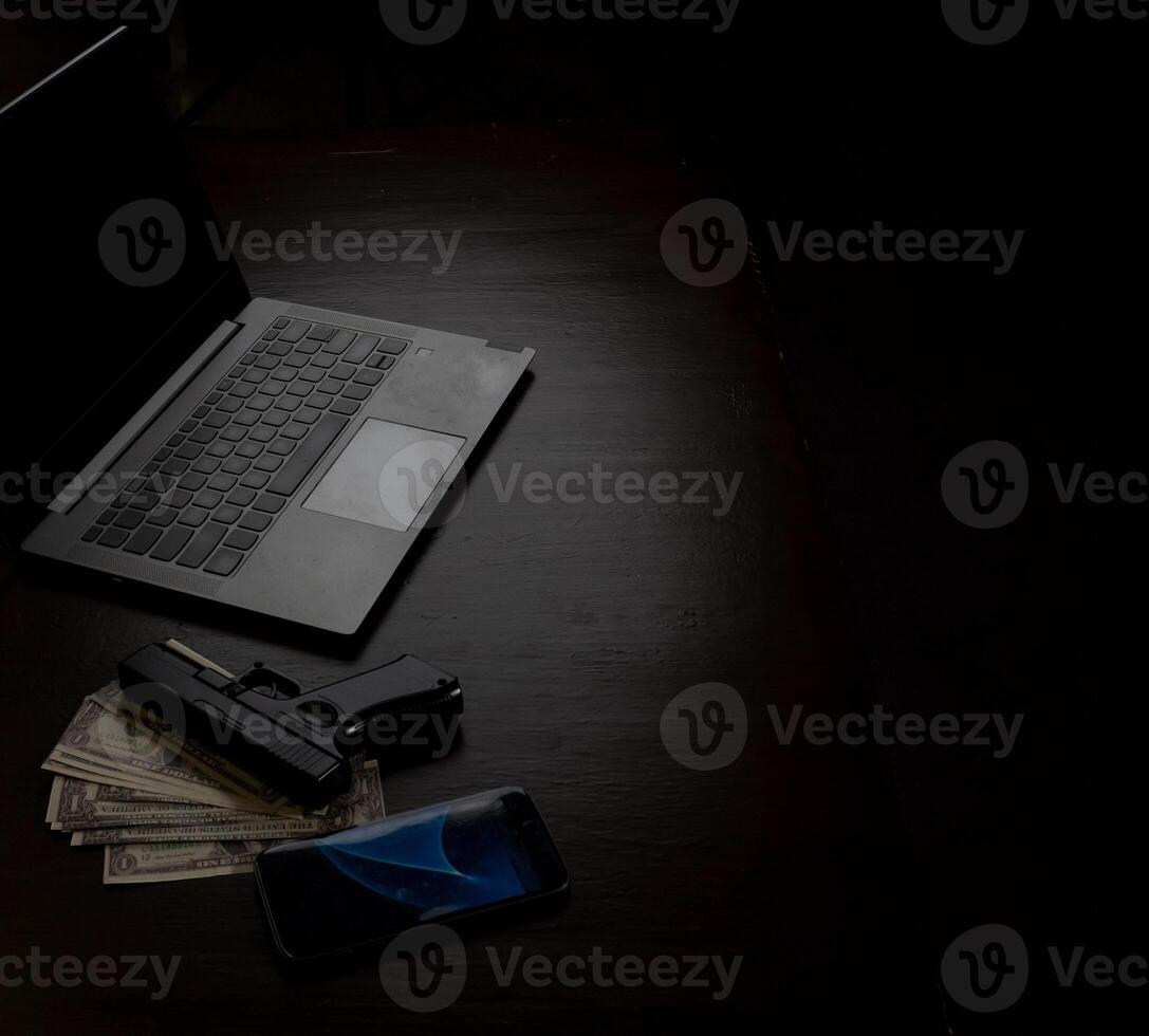 Computers, mobile phone, money, guns, and other devices that hackers use to steal or hijack information to trick victims into taking money. laying on wooden table in dark room in mysterious apartment. photo