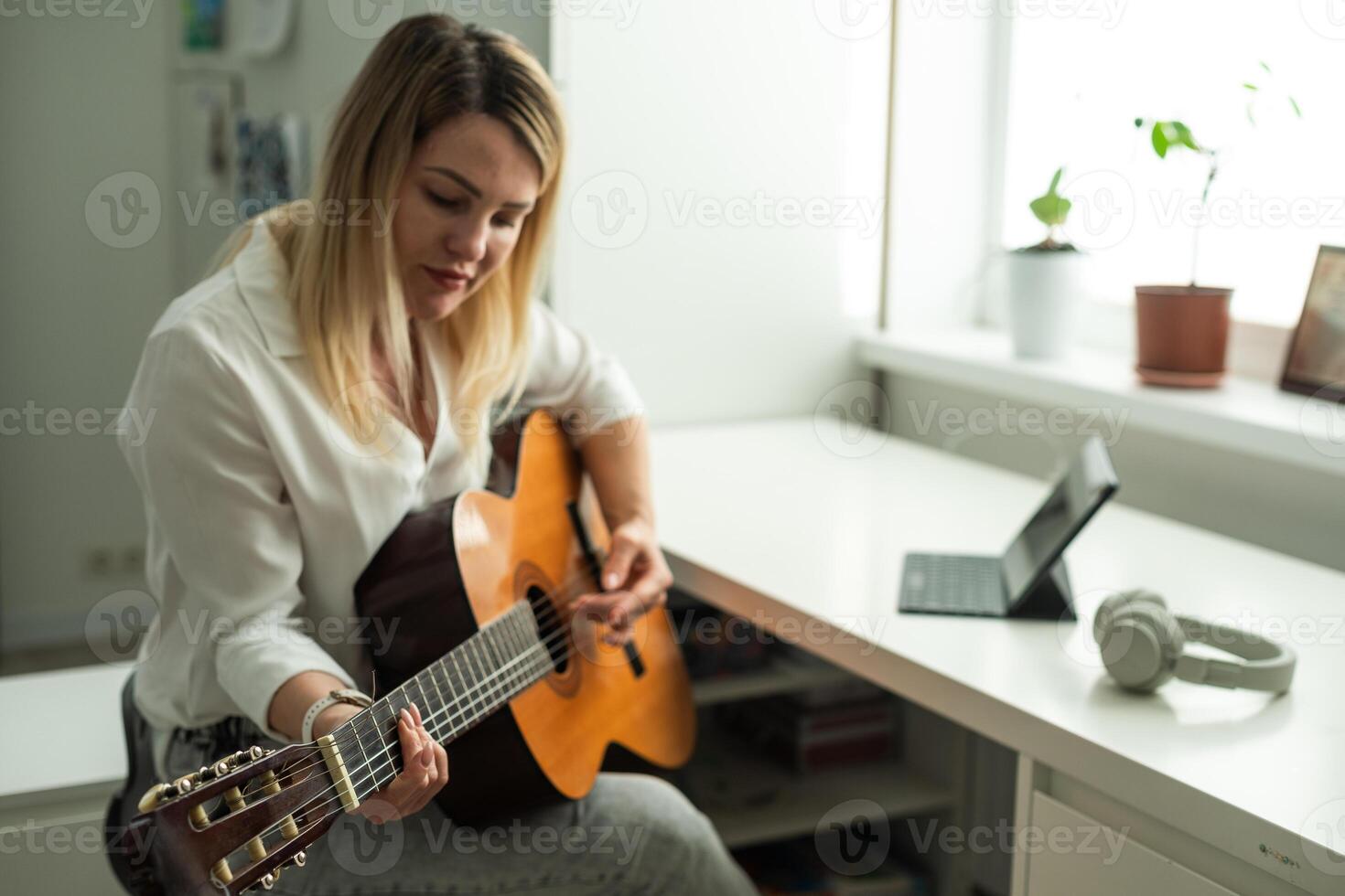 Home Guitar Playing Lesson Girl Hold Instrument. Beautiful Woman at Musical Training. Blonde with Makeup Looking at Notes on Music Stand. Caucasian Girl Guitarist Taking Chords on Strings photo