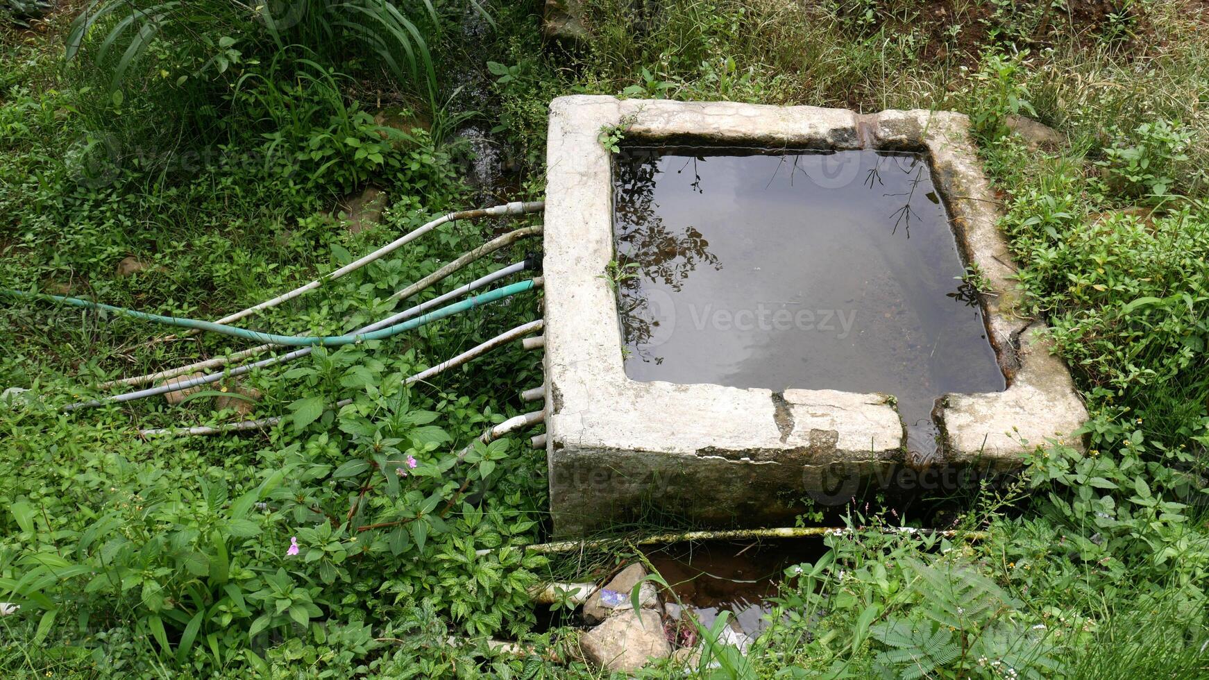 Irrigation system in the countryside with concrete water tank as a water storage, with some plastic pipes used for distribution to farms or households. Wild plants grow abundantly around it. photo
