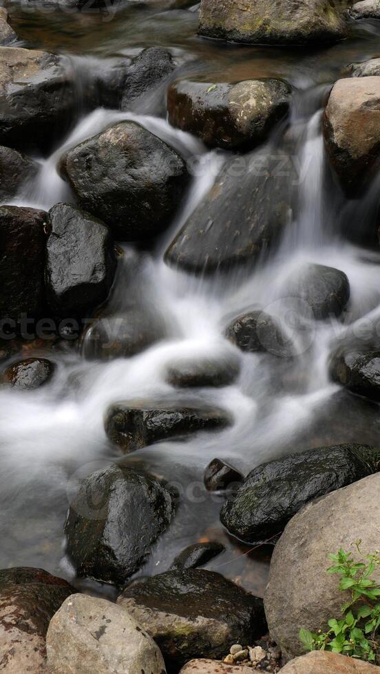 Long Exposure, clear river water flows between large rocks, offering a serene view with green foliage adorning the riverbanks. photo