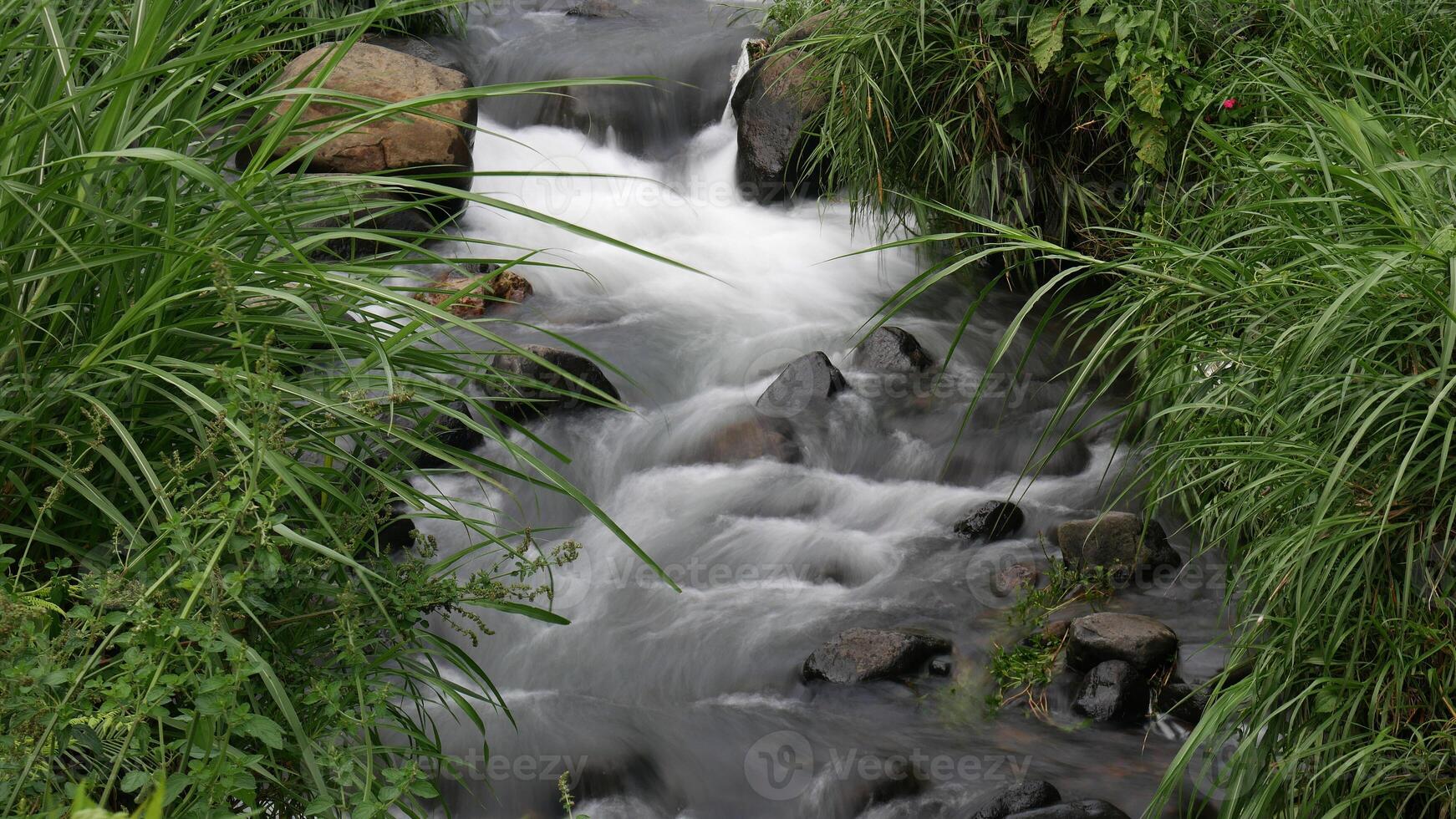 Long exposure calm stream flows between large rocks, creating a peaceful and cool atmosphere, with touches of green foliage around the stream. photo