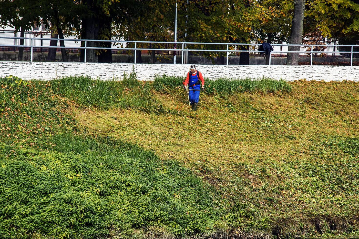 Nitra, Slovakia - 11.18.2024 A municipal worker cuts the grass using power tool string lawn trimmer on the banks of the Nitra river. photo