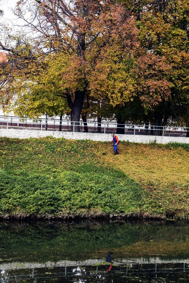 Nitra, Slovakia - 11.18.2024 A municipal worker cuts the grass using power tool string lawn trimmer on the banks of the Nitra river. photo