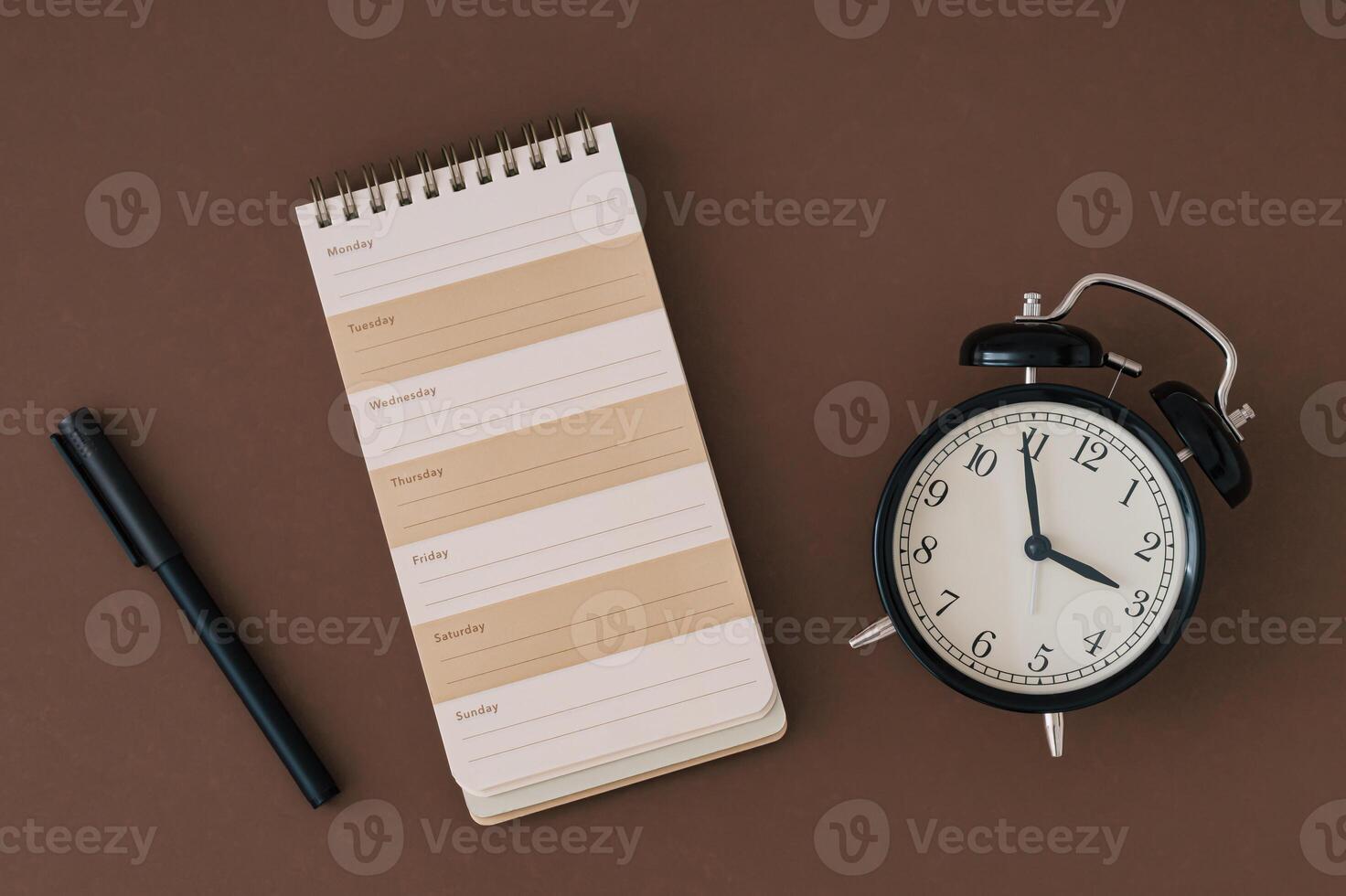 Planner pad, alarm clock and pen on brown background. Planner for organization and goal priorities. Writing down tasks and schedule reminders on office desk. Trendy brown color flat lay. photo