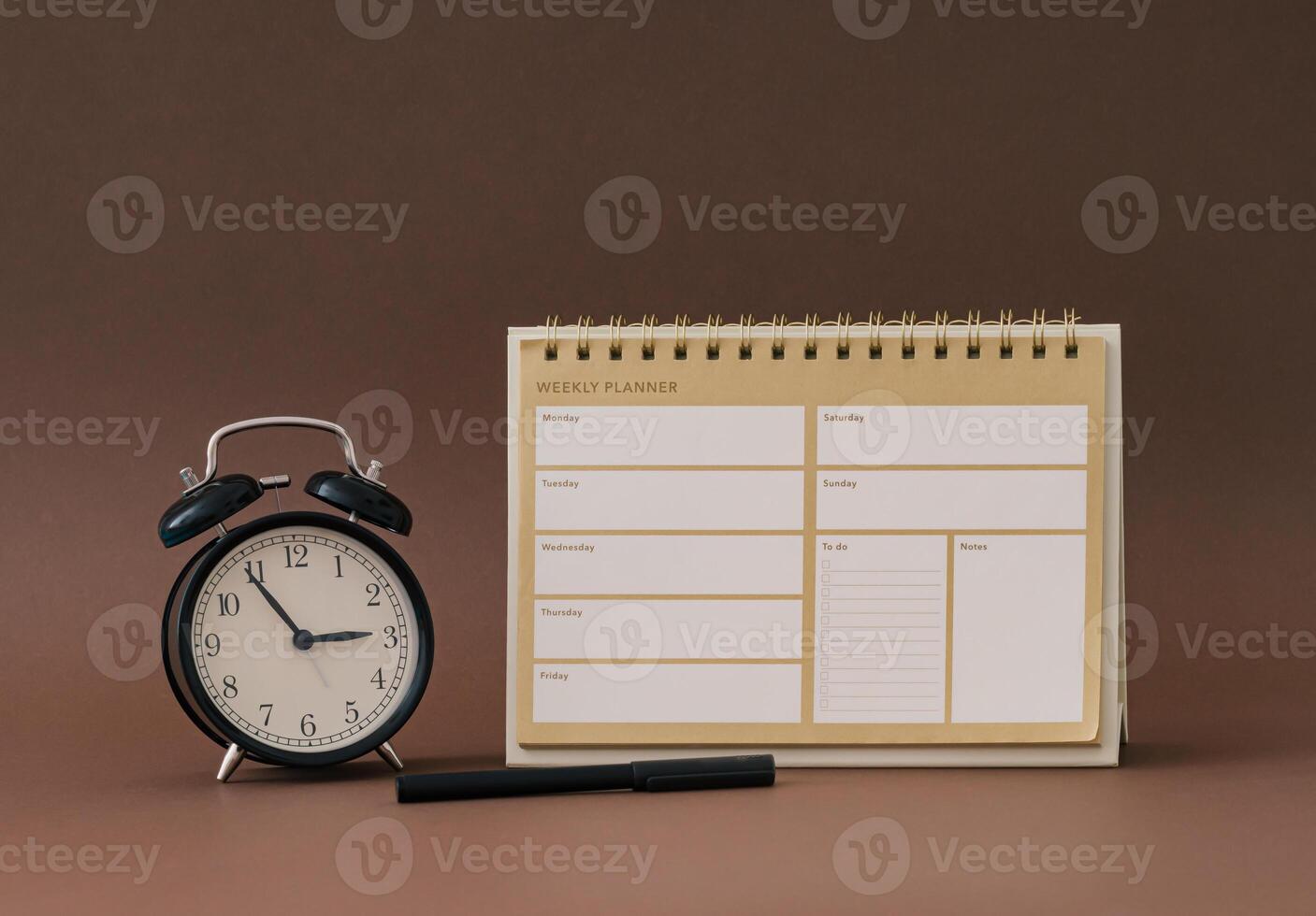 Planner pad, alarm clock and pen on brown background. Planner for organization and goal priorities. Writing down tasks and schedule reminders on office desk. Trendy brown color layout. photo