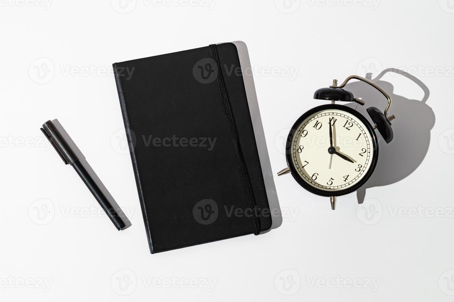 Black notebook, alarm clock and pen on white background. Planner for organization and goal priorities. Writing down tasks and schedule reminders on office desk. Minimal creative concept. Flat lay. photo