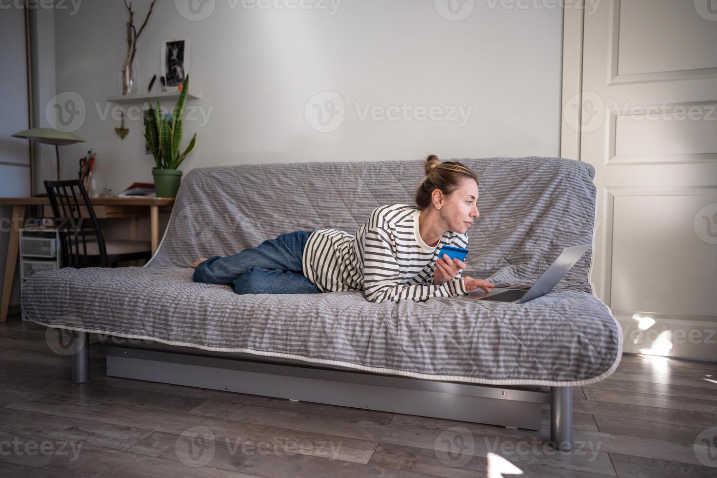 Relaxed online shopping of woman while comfortable lying with banking card on couch, using laptop. photo
