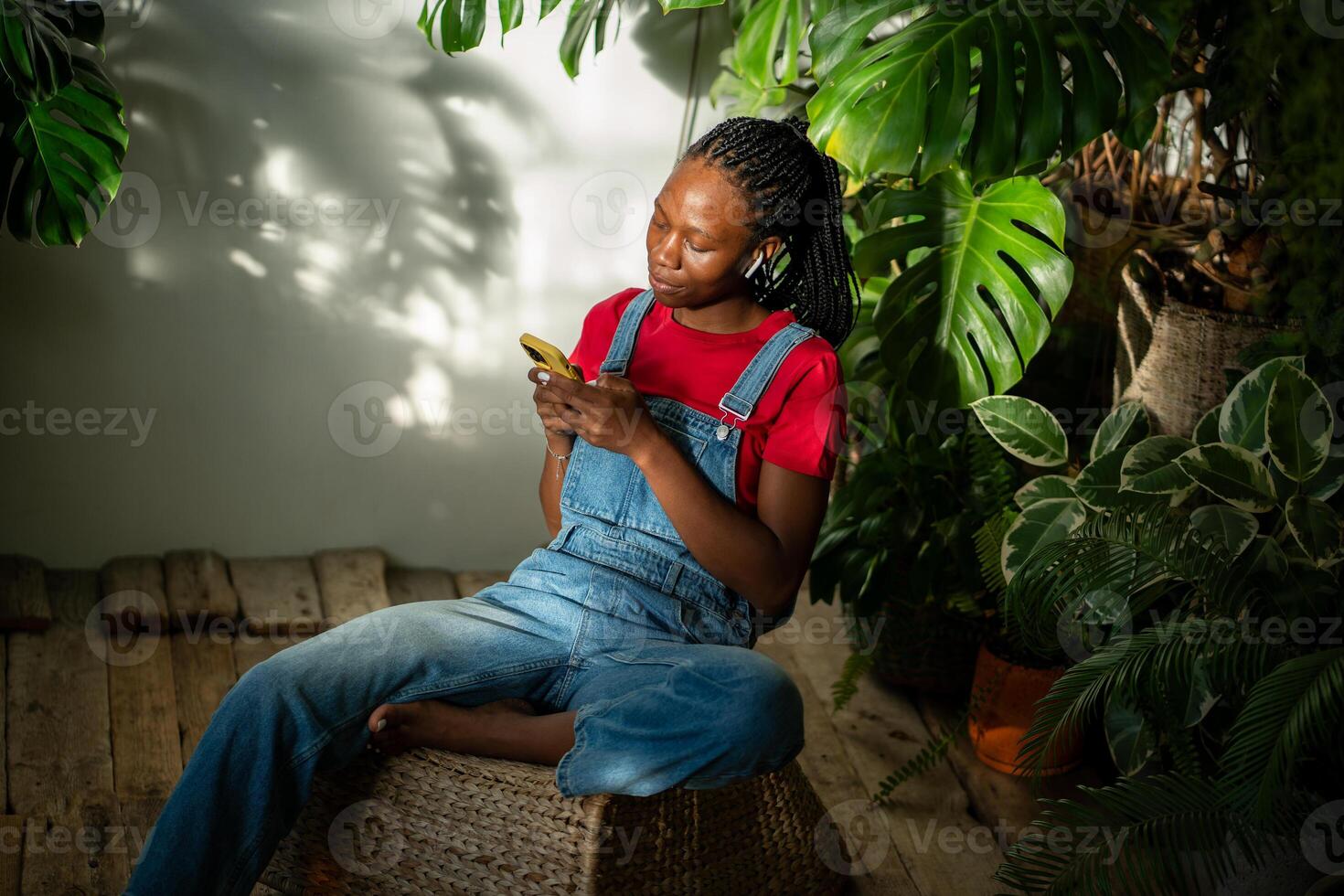 Relaxed black girl in earphones holds smartphone typing message, listen music in room with monstera. photo