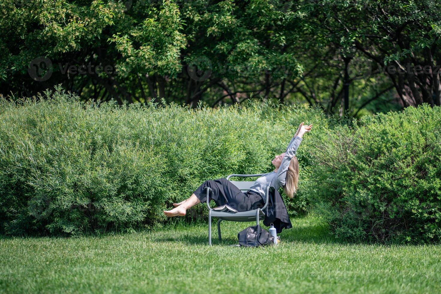 Carefree woman relaxing on park chair surround lush plants barefoot stretching arms on break outdoor photo