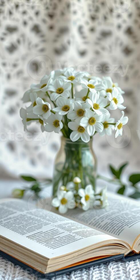 Fresh white flowers in a glass jar set on an open book against a soft patterned background photo