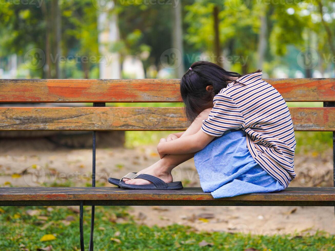 A sad little girl is sitting alone on bench in park. The little girl is unhappy and anxious because she has no one to play with. photo