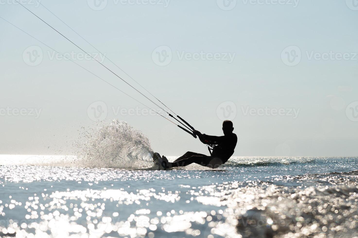 Kite surfing on waves. Silhouette of athletic man riding fast on kiteboard in sea, creating waves and splashes on water photo