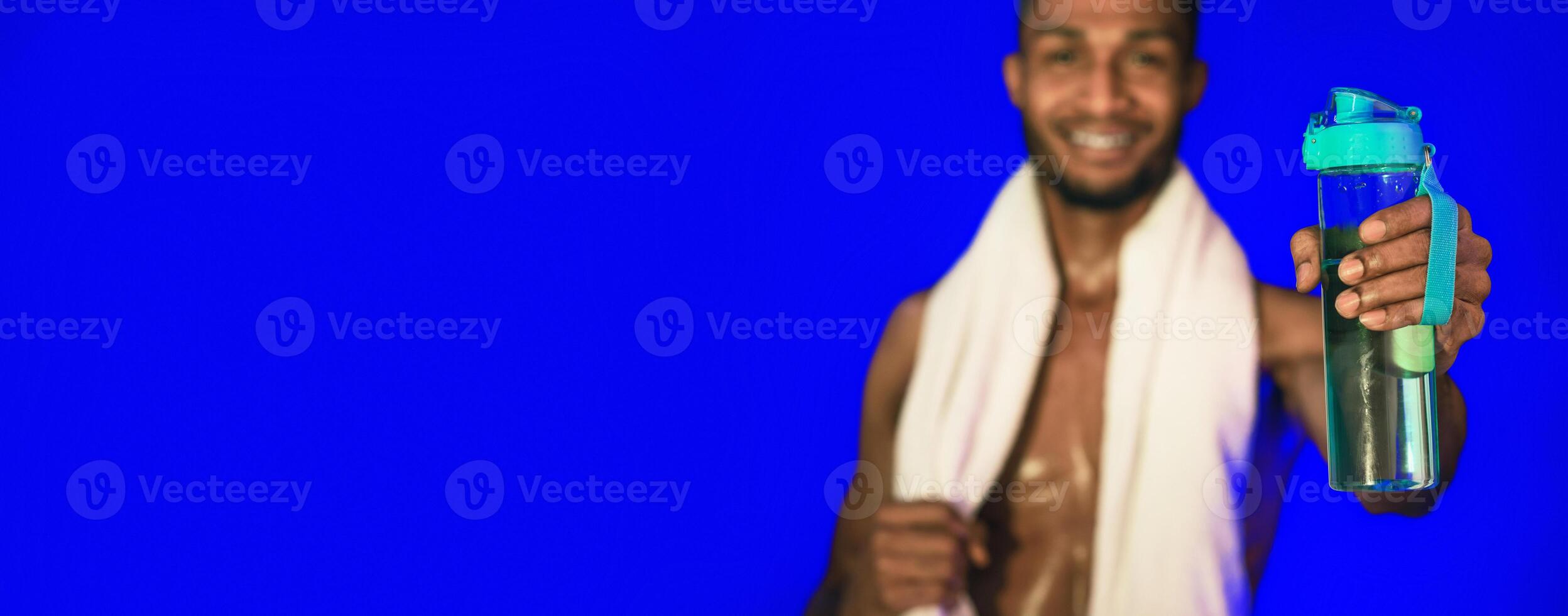 Workout Hydration. Black Athlete Man Offering Bottle Of Water To Camera Posing On Blue Studio Background. Shallow Depth, Selective Focus photo