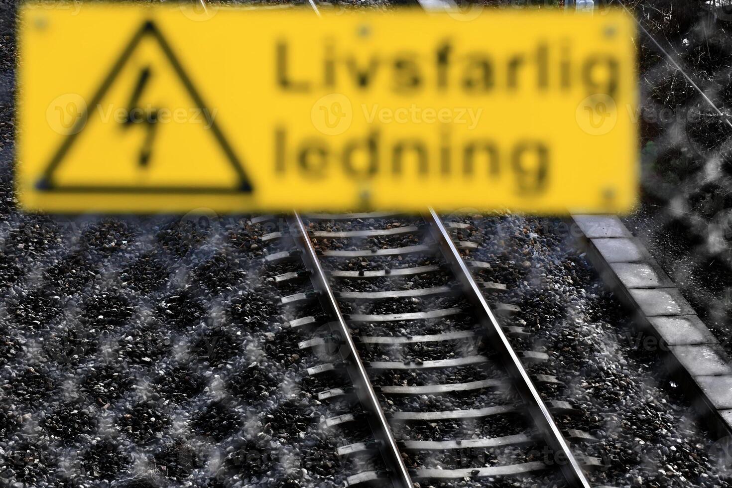 Defocused fence and warning sign in front of rail track. The sign says Lifsfarlig ledning - Cable dangerous to life - in Swedish photo